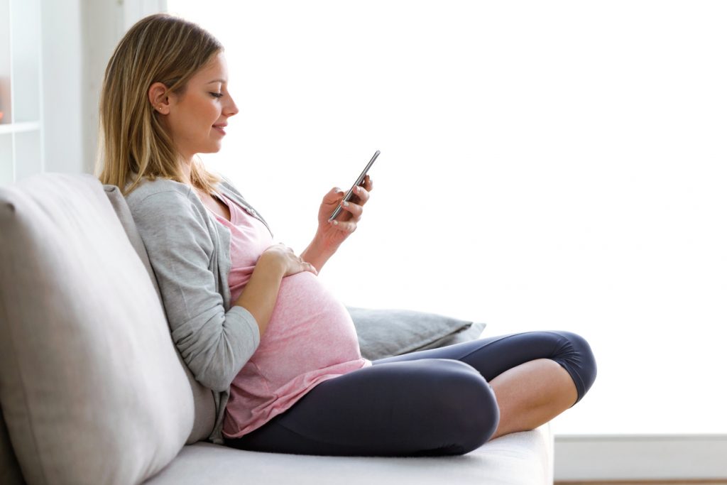 Beautiful young pregnant woman texting with her smartphone on the sofa at home